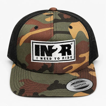 A camouflage trucker hat with a black mesh back and a front patch reading 'IN2R I NEED TO RIDE'. A circular 'The Authentic Snapback' sticker is visible on the brim.