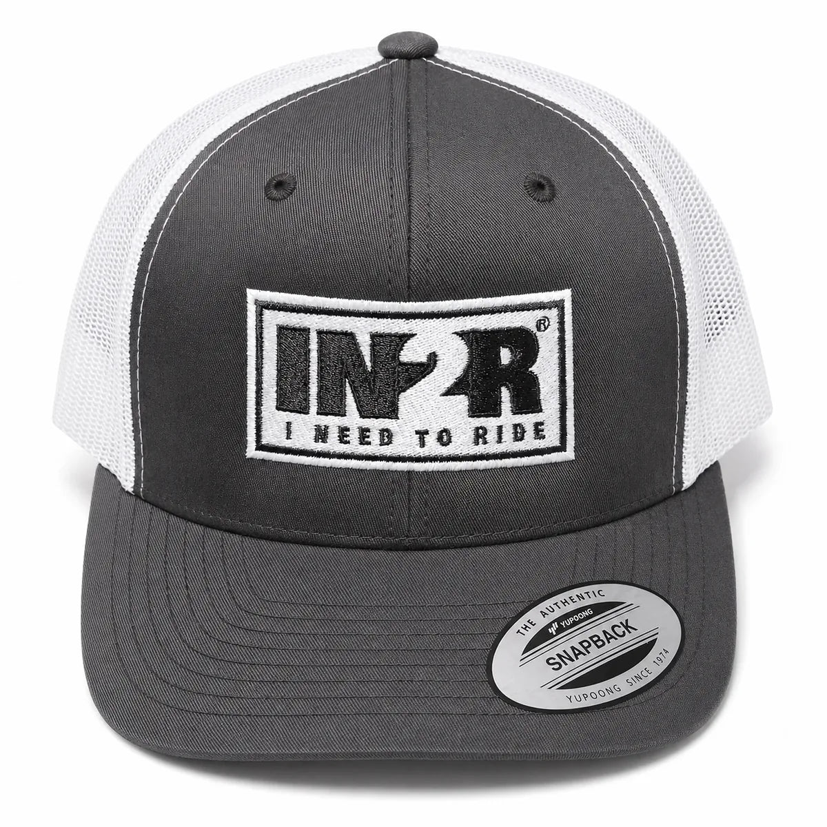 A dark gray and white trucker hat with a black and white embroidered front patch reading 'IN2R I NEED TO RIDE'. A circular sticker on the brim reads 'The Authentic Snapback'.
