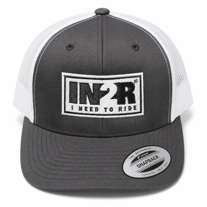 A dark gray and white trucker hat with a black and white embroidered front patch reading 'IN2R I NEED TO RIDE'. A circular sticker on the brim reads 'The Authentic Snapback'.
