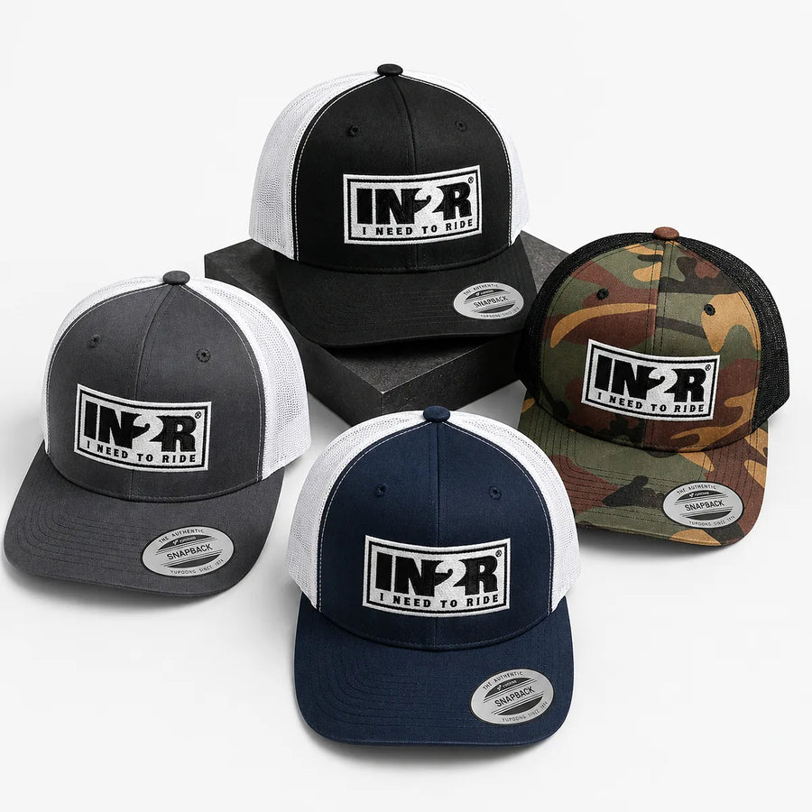 Four trucker-style snapback caps arranged in a 2x2 stack on a wooden surface, with a blurred outdoor mountain background. The caps are shown in four colorways: black and white, camouflage, dark gray and white, and navy blue and white.