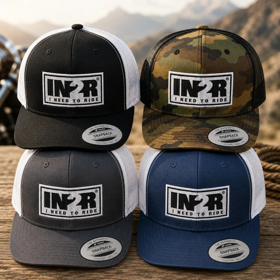Four trucker-style snapback caps arranged in a 2x2 stack on a wooden surface, with a blurred outdoor mountain background. The caps are shown in four colorways: black and white, camouflage, dark gray and white, and navy blue and white.