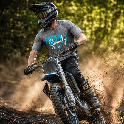 A person riding a dirt bike on a muddy forest trail, wearing a black helmet, gray t-shirt with blue and white 'Simply Gnarly' text, and black pants. Dust is kicked up behind the bike.