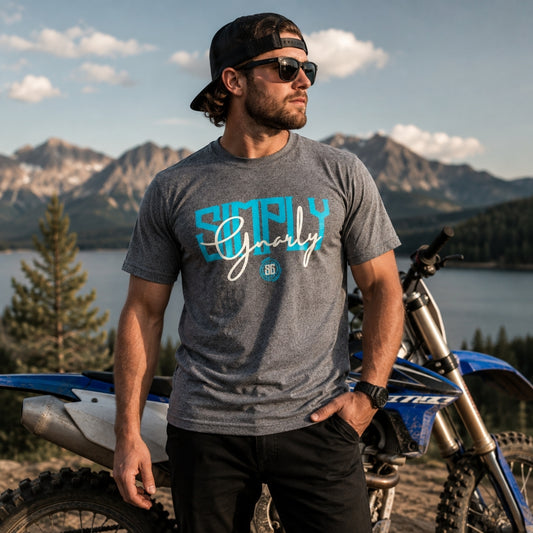 A man wearing a gray heather t-shirt with a blue and white 'Simply Gnarly' graphic stands in front of a blue and white dirt bike, with a mountain lake landscape in the background.