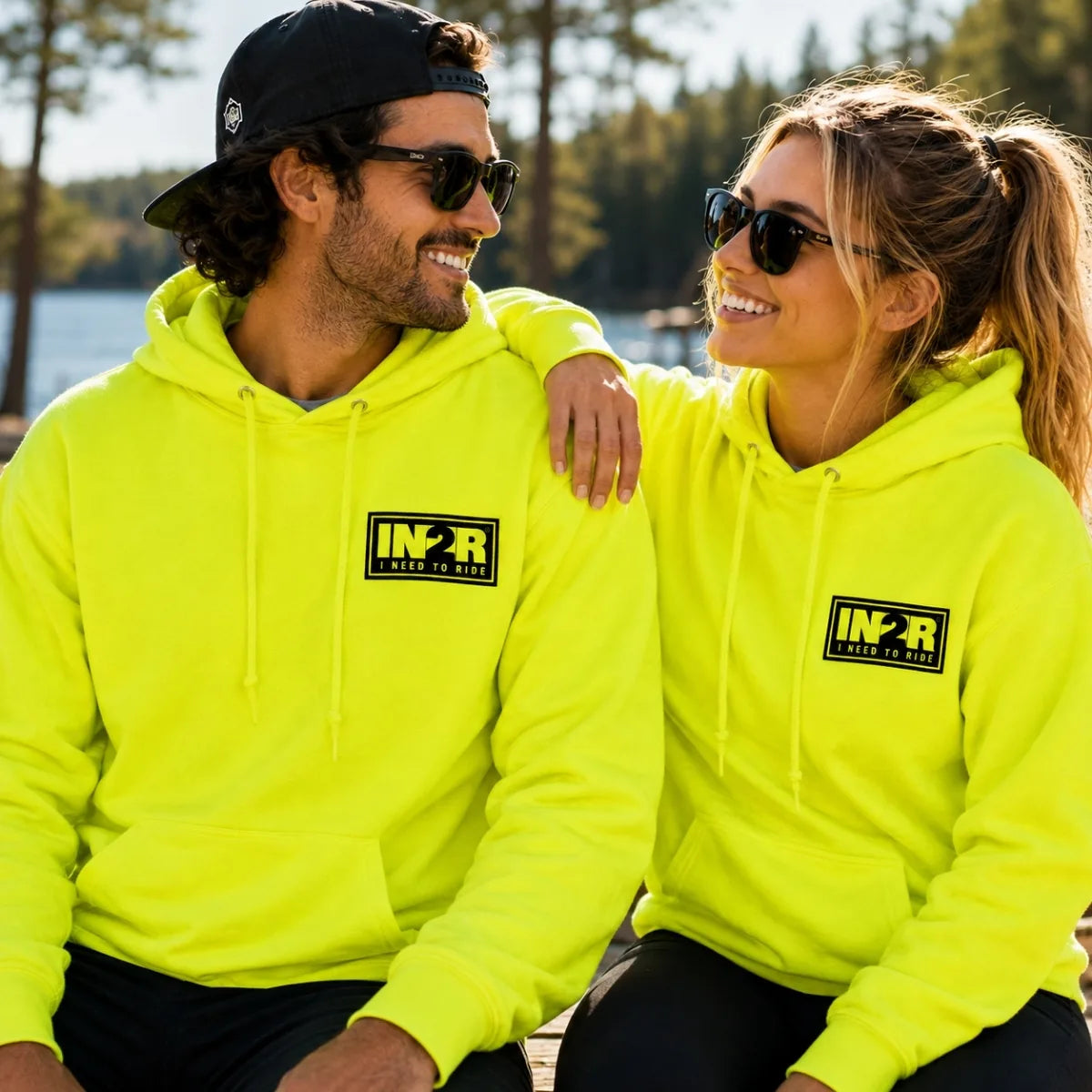 A man and a woman wearing matching neon yellow-green hoodies with a black and yellow 'IN2R' logo on the chest, sitting outdoors near water and trees.