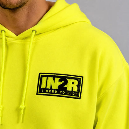A close-up of a person wearing a bright neon yellow hoodie with a black and yellow 'IN2R I NEED TO RIDE' logo patch on the chest, set against a neutral gray background.