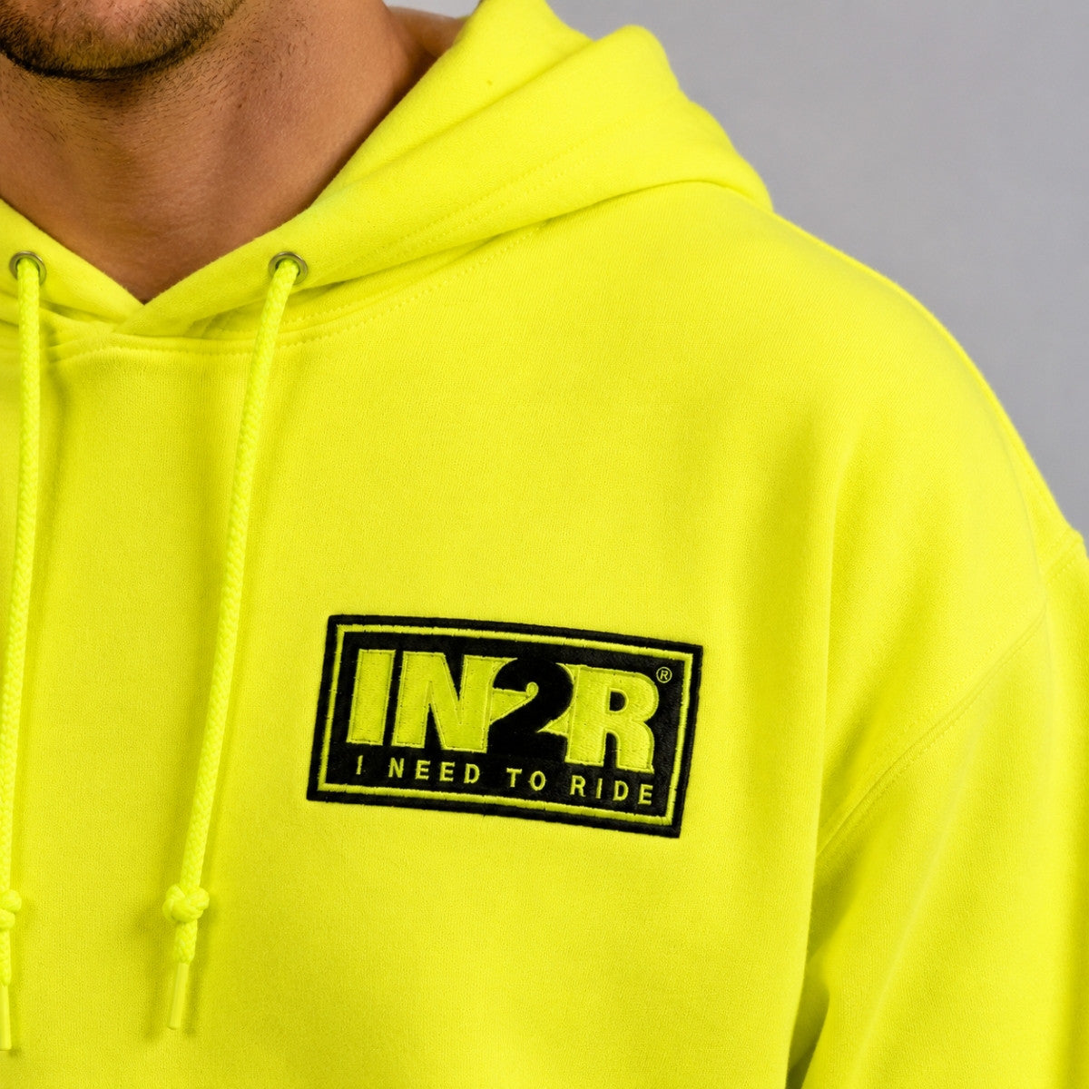 A close-up of a person wearing a bright neon yellow hoodie with a black and yellow 'IN2R I NEED TO RIDE' logo patch on the chest, set against a neutral gray background.