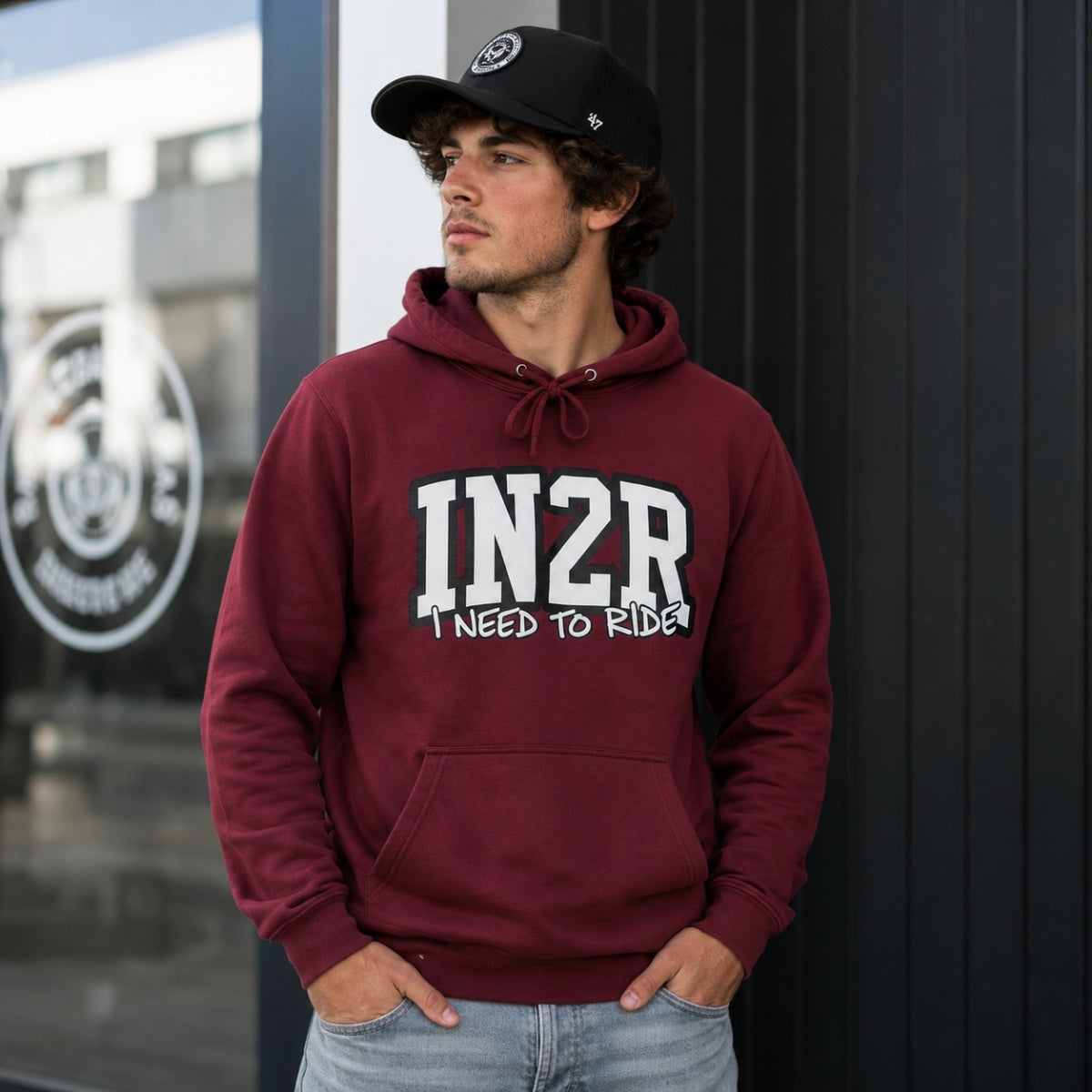A man wearing a maroon hoodie with 'IN2R I NEED TO RIDE!' printed in white on the front, paired with a black baseball cap and light blue jeans, standing against a dark paneled wall.