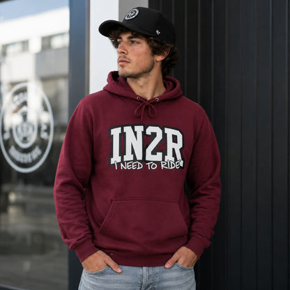 A man wearing a maroon hoodie with 'IN2R I NEED TO RIDE!' printed in white on the front, paired with a black baseball cap and light blue jeans, standing against a dark paneled wall.