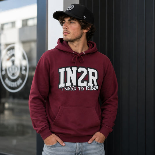 A man wearing a maroon hoodie with 'IN2R I NEED TO RIDE!' printed in white on the front, paired with a black baseball cap and light blue jeans, standing against a dark paneled wall.