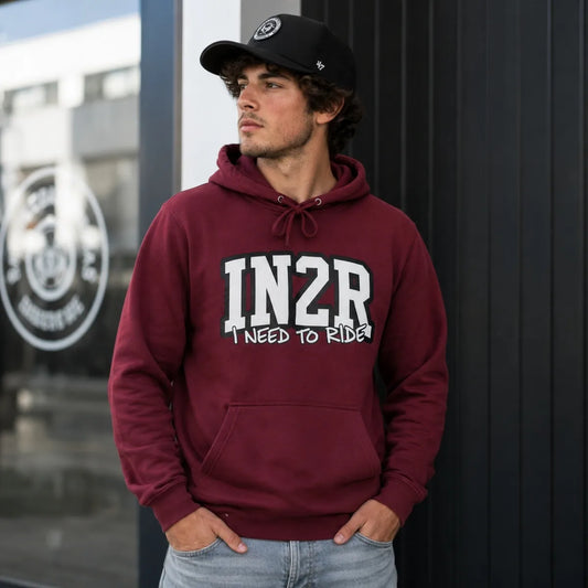 A man wearing a maroon hoodie with 'IN2R I NEED TO RIDE!' printed in white on the front, paired with a black baseball cap and light blue jeans, standing against a dark paneled wall.