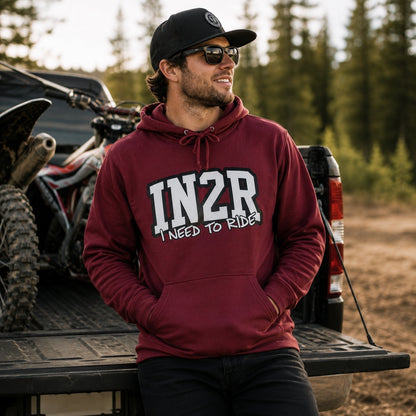 A man wearing a maroon hoodie with '1N2R I NEED TO RIDE' printed on the front, sitting in the bed of a pickup truck with an ATV in the background.
