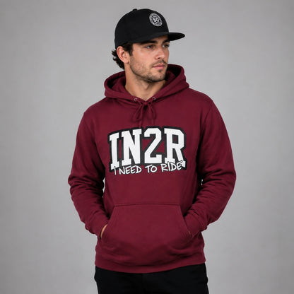 A man wearing a maroon pullover hoodie with 'IN2R I NEED TO RIDE' printed in white on the front, paired with a black baseball cap, set against a plain gray studio background.