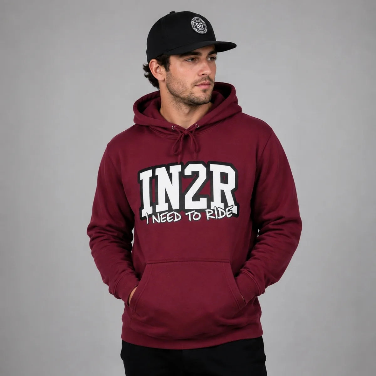 A man wearing a maroon pullover hoodie with 'IN2R I NEED TO RIDE' printed in white on the front, paired with a black baseball cap, set against a plain gray studio background.