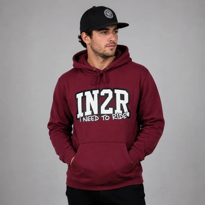 A man wearing a maroon pullover hoodie with 'IN2R I NEED TO RIDE' printed in white on the front, paired with a black baseball cap, set against a plain gray studio background.