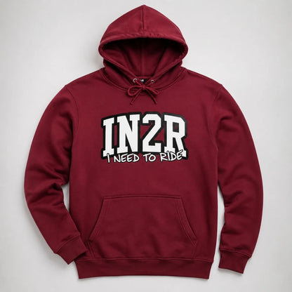 A maroon pullover hoodie with a front kangaroo pocket and drawstring hood, featuring the text 'IN2R' in large white varsity letters with black outlines and 'I NEED TO RIDE!' underneath, shown on a light gray background.