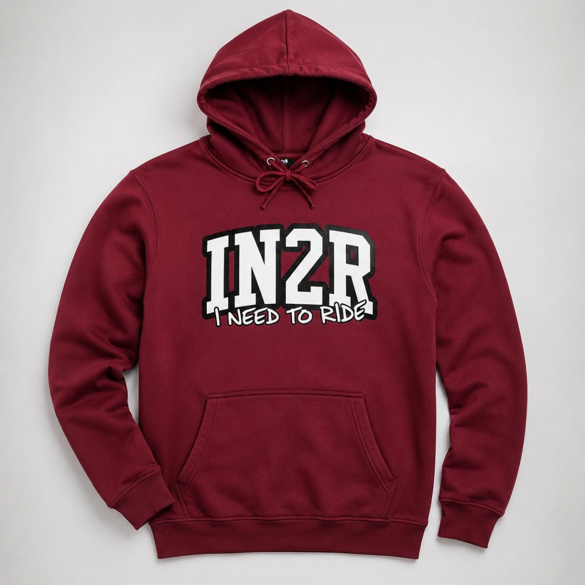 A maroon pullover hoodie with a front kangaroo pocket and drawstring hood, featuring the text '1N2R' in large white varsity letters with black outlines and 'I NEED TO RIDE!' underneath, shown on a light gray background.