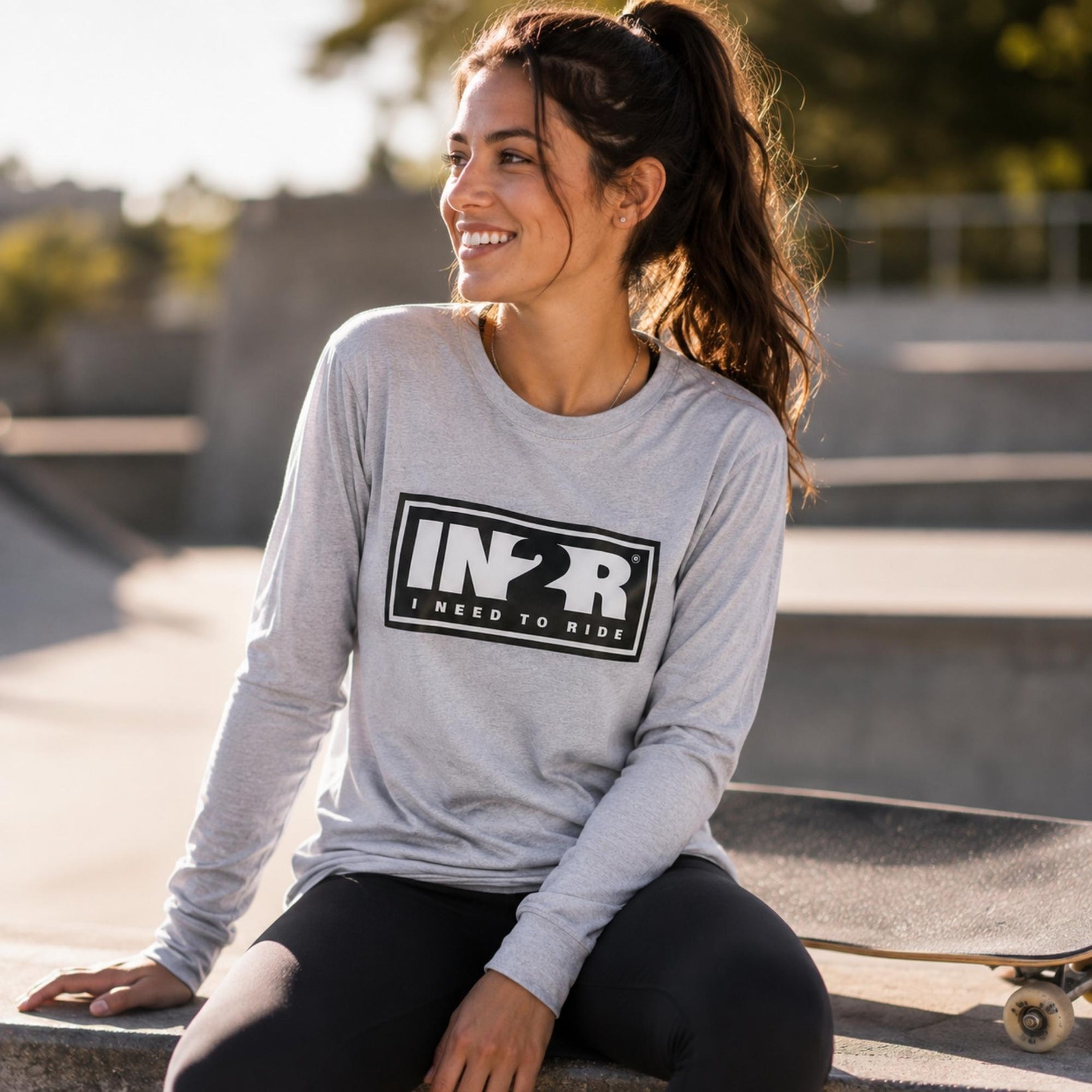 A woman sitting outdoors at a skatepark wearing a light gray long-sleeve t-shirt with a black 'IN2R I NEED TO RIDE' graphic on the front, paired with black leggings.