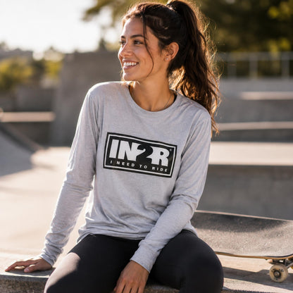 A woman sitting outdoors at a skatepark wearing a light gray long-sleeve t-shirt with a black 'IN2R I NEED TO RIDE' graphic on the front, paired with black leggings.