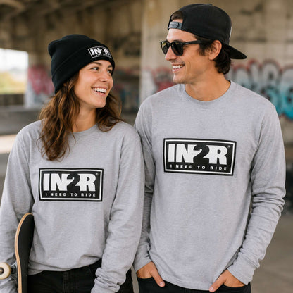 A man and a woman wearing matching heather gray long-sleeve crewneck shirts with a black and white 'IN2R I NEED TO RIDE' graphic on the chest, standing in an outdoor urban setting.
