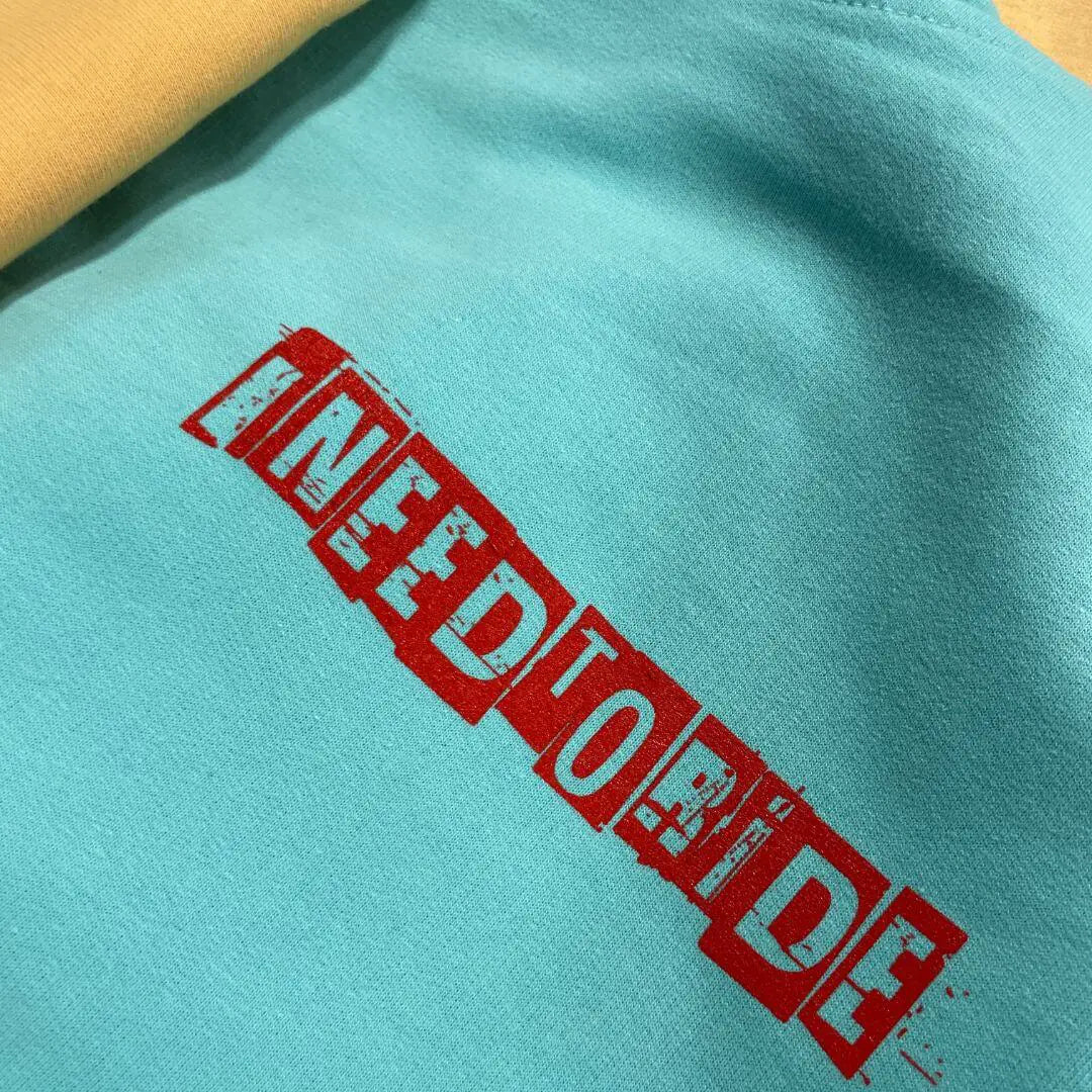 A close-up of a mint colored fabric, on the front of hoodie, featuring a bold red distressed graphic that reads 'I NEED TO RIDE' printed diagonally across the chest.