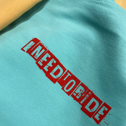 A close-up of a mint colored fabric, on the front of hoodie, featuring a bold red distressed graphic that reads 'I NEED TO RIDE' printed diagonally across the chest.