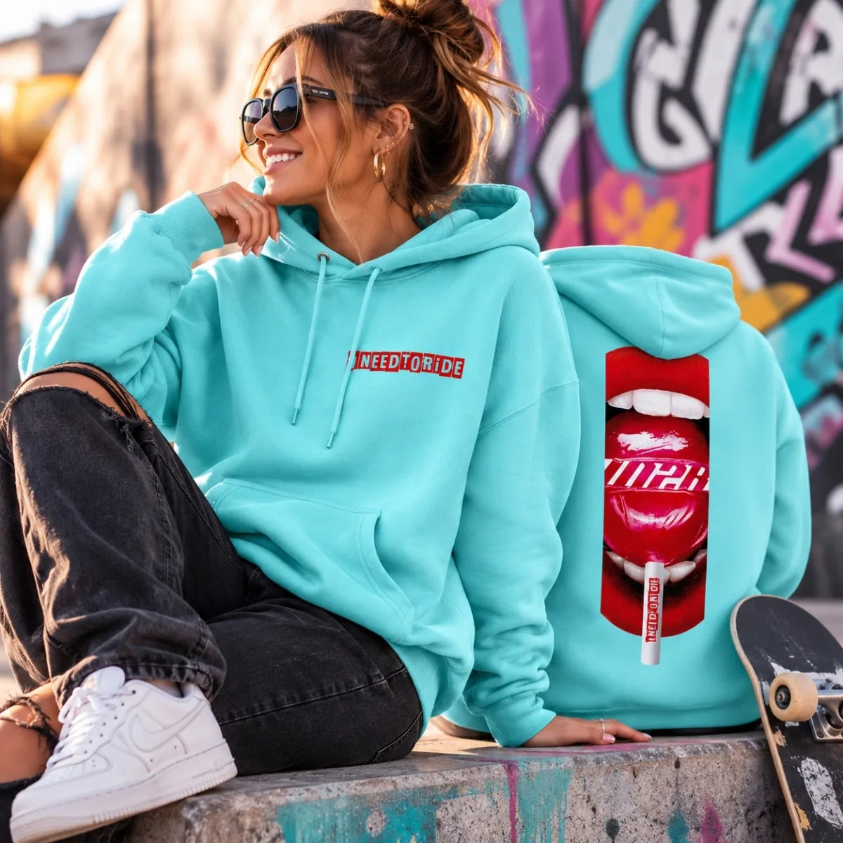 A woman wearing a mint colored hoodie sits on a concrete ledge in an outdoor urban setting with graffiti walls behind her. The hoodie has a red rectangular chest logo reading 'I NEED TO RIDE' and a large red lips graphic on the back with 'IN2R' and a small red-and-white tube graphic.